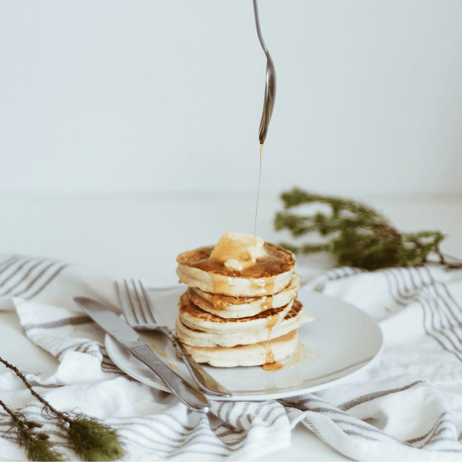 Stapel pannenkoeken met een klontje boter en siroop, geserveerd op een wit bord met bestek.
